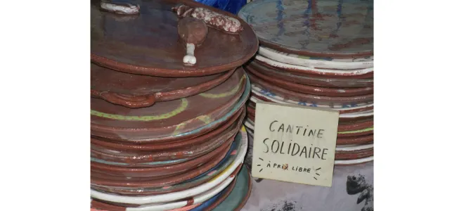 Piles d’assiettes en céramique colorée avec un panneau manuscrit « Cantine solidaire à prix libre », symbole de convivialité et de partage à Uzerche.