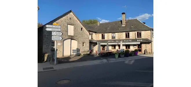 Façade de l'Hôtel de la Poste à Saint-Privat, bar-restaurant de village en pierre avec terrasse fleurie, panneaux directionnels et ciel bleu.