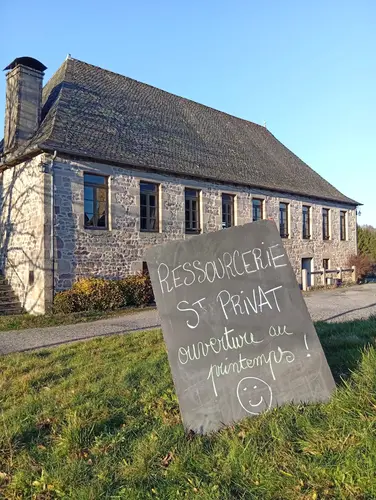 Ancienne salle des fêtes de Saint-Privat, futur local de la ressourcerie, avec un panneau manuscrit "Ressourcerie St Privat, ouverture aux Printemps !" devant le bâtiment en pierre.