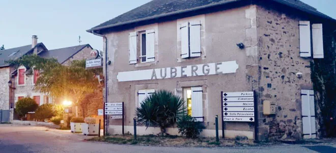 L'image montre l'extérieur de l'Auberge de la Tanière, illuminée à la tombée de la nuit, symbolisant un lieu communautaire et culturel revitalisé par trois amis dynamiques dans la petite commune d'Orliac-de-Bar. Ce lieu, autrefois un restaurant et un hébergement menacé de fermeture, est devenu un espace de lien social, proposant des activités variées comme des projections et des tournois, tout en servant de cantine pour l'école locale.
