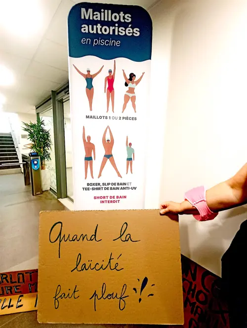 Affiche officielle "Maillots autorisés en piscine" du Centre Aqua de Tulle montrant des silhouettes de maillots de bain autorisés, devant laquelle une main tient une pancarte écrite à la main : "Quand la laïcité fait plouf !" – illustration de la mobilisation contre l'interdiction des maillots couvrants.