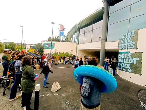 Photo d'une mobilisation du collectif MCMMMC devant l'entrée du Centre Aqua à Tulle : un groupe de manifestants avec des banderoles, une personne portant une grande bouée bleue autour du cou, et sur le mur l'inscription "Mon corps mon maillot mon choix".